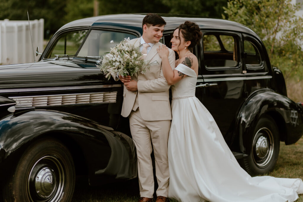 A bride and groom look at each other while leaning against a classic 
vintage car.