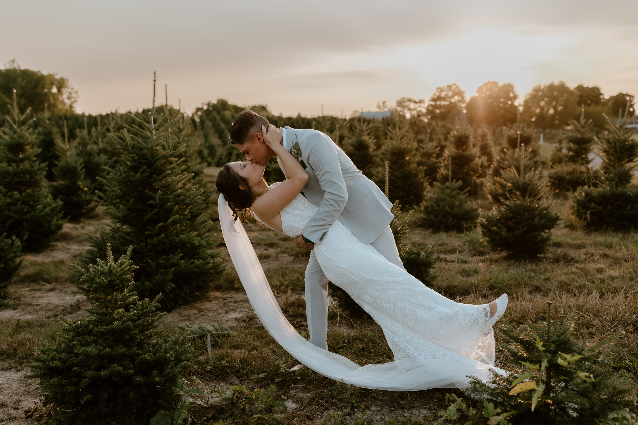 Groom dips bride and kisses her during golden hour in a pine tree forest